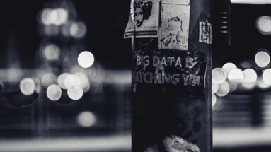 A telephone pole covered in stickers, including a large one that reads "BIG DATA IS WATCHING YOU"
